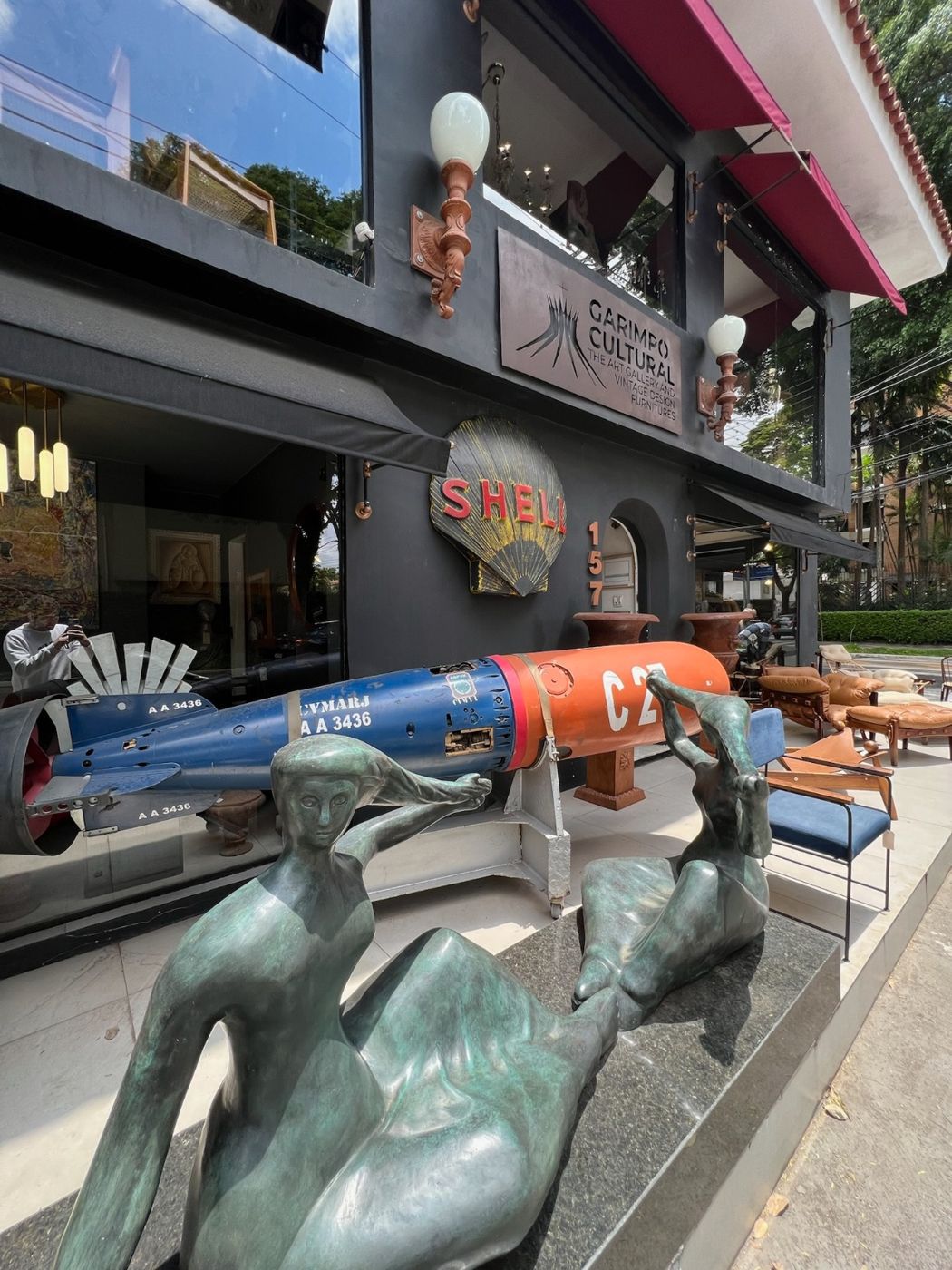 Fachada da Garimpo Cultural, com esculturas em bronze, um torpedo antigo e poltronas de couro na calçada